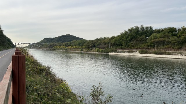 A river scene with a smooth water surface, flanked by a cycling path and vegetation. In the background, there’s a bridge over the river, and hills with on the other side.