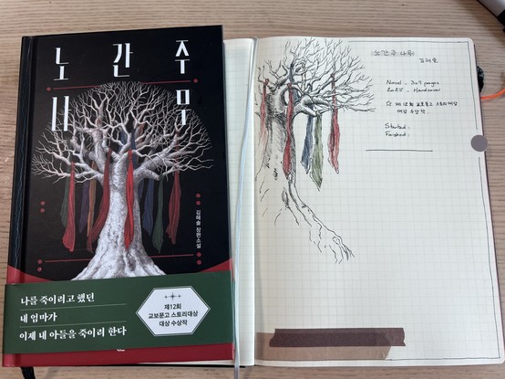 An opened notebook and a Korean hardcover book with the illustration of a leafless tree with coloured ribbons attached to its branches. The page of the notebook shows a drawing of the said tree and handwritten information about the book. 