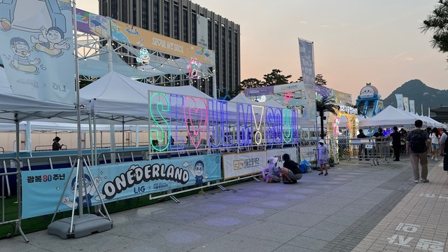 An outdoor event area featuring colorful signage reading "Seoul My Soul." The scene includes white tents, promotional banners, and swimming pools barely visible behind the banners. Mountain and building in the distance. 