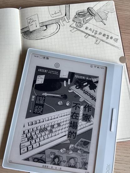An e-reader and a notebook on a table. The e-reader displays a book cover with Chinese text, a desktop set-up with keyboard, computer screen, magazine, bottle of ink, etc. The notebook shows the reproduction with pen and pencil of some elements of the cover.