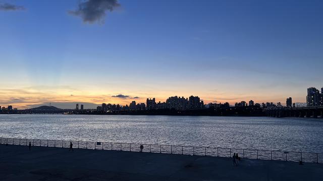 A sunset landscape focused on a city outline across a large river. The buildings are black against a sky turning from dark orange to dark blue. 