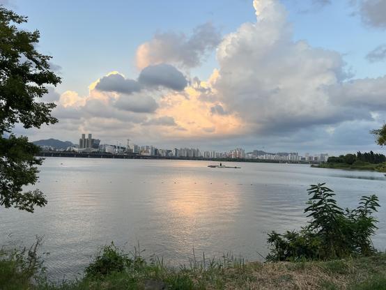 A large river with a city skyline on the other bank and a small boat in the distance. The sky takes half of the picture and has big clouds with different shades of pink and grey. 
