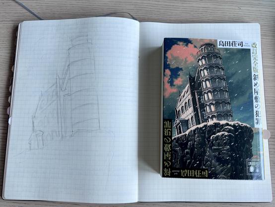 A Japanese pocket book resting on the right page of a grid notebook. The cover shows a dramatic illustration of a mansion attached to a tower on a rocky cliff. The building is leaning to the left. The left page of the notebook shows a pencil sketch of the same building. 