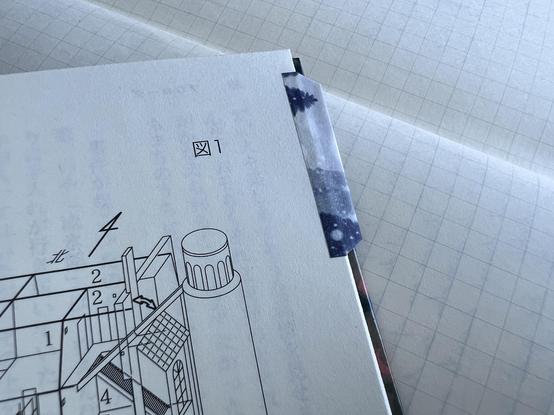 Top corner of a book opened on a page  showing the plan of a mansion with several floors and rooms. At the top of the page, a piece of washi tape has been folded and cut to stick out of the page and serve as bookmark. 