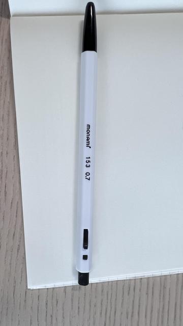 Focus of the picture is a retractable ballpoint pen with a hexagonal white body in plastic, a black grip and a black button to release the tip. “Monami 153 0.7” is written on the body. The pen looks simple and cheap. 