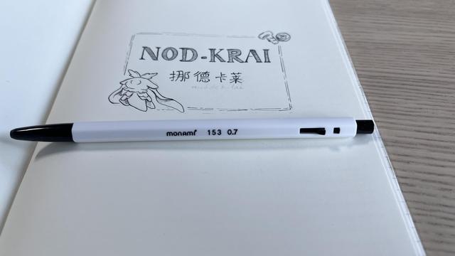An open notebook page featuring the word “Nod-Krai” in English and Chinese inside a rectangular frame. A small magical-looking creature and two small screws complete the frame. A retractable ballpoint pen is lying on the page.