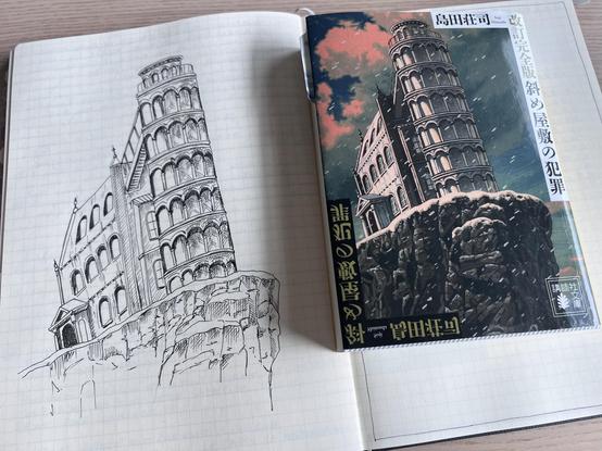 A book in Japanese on a notebook. The book cover shows a mansion with a tower that’s leaning to its side on a rocky mountain. This illustration has been (poorly) reproduced on the notebook. 