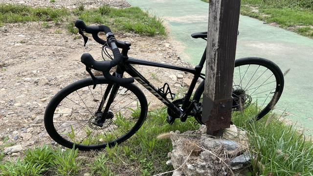 A black road bike is leaning against a wooden post, with grass and dirt around. A path is visible on the back. 