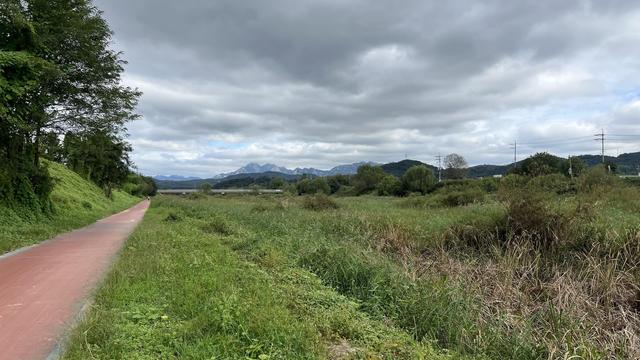 Outdoor scenery with a red path on the left and a vast area of grass and plants on the right. Mountains are visible in the distance and heavy grey clouds cover the sky. 