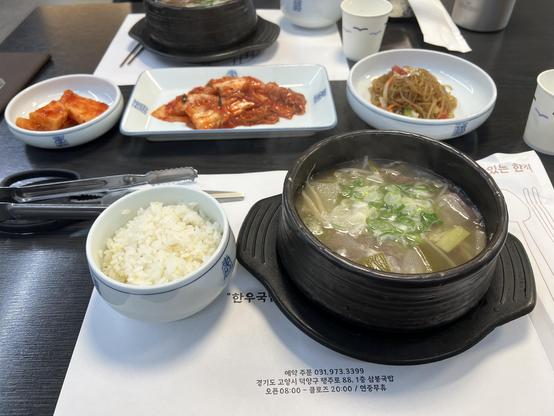 Table in a restaurant with a hot pot containing a light soup, a bowl of rice and various side dishes. 