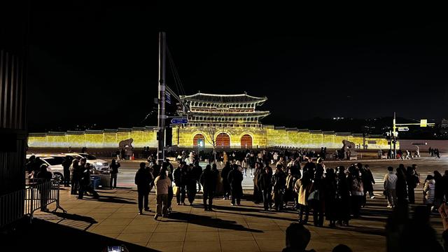 Picture taken by night of a long stone wall and a huge traditional Korean gate at the center with three red arches and a double roof pavilion. The whole façade is illuminated with lights. The sky is black and a crowd of people is on the foreground, waiting to cross a street. 