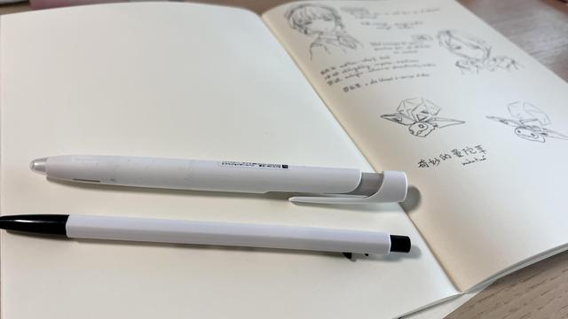 A notebook opened. The left page is blank with two white pens on it, one is very thin and the other looks more ergonomic. The right page is out of focus but is filled with doodles and notes. 
