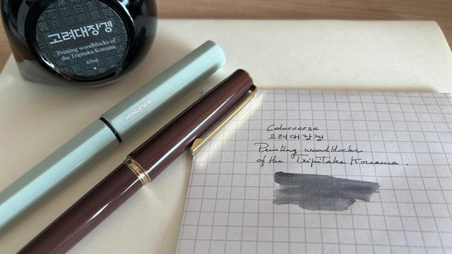Picture of two fountain pens on a desk, with a bottle of dark ink on the top left of the picture (partly outside the frame). The label is written in Korean. Near the pens, there is a small sheet of paper with the name of the ink handwritten and an ink swab on it. 