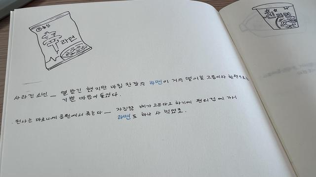 Page of a blank notebook. Focus is on the left page where there’s a doodle of ramyeon package and two quotes in Korean, both with the word “ramyeon” written in blue. The doodle of a ramyeon  cup is on top of the right page.