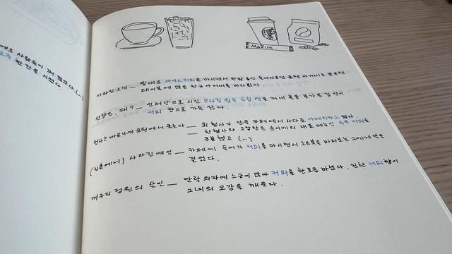An opened blank notebook displays a page with doodles of coffee on top (cup of hot coffee, glass of iced americano, instant coffee, coffee bean bag…) and several handwritten lines in Korean with some words highlighted in blue. 