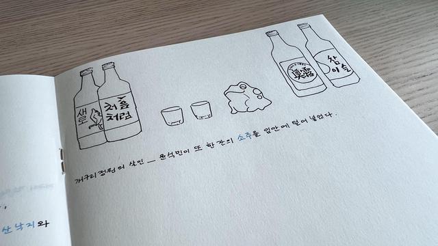 Top half of a notebook page with doodles at the top: four bottles of soju from various brands, two soju glasses and a small frog. Below the doodles, there is a line of text in Korean with the word for soju written in blue. 