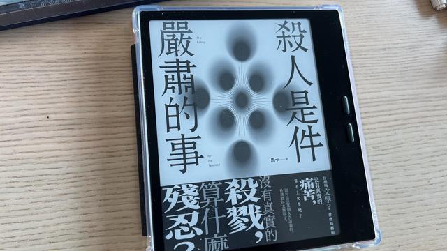 An e-reader on a wooden table displaying a book cover in black and white. The title is 殺人事件嚴肅的事, the cover is white with dark dots in the middle and there is a promotional wrapping band printed on the cover.