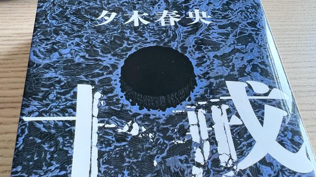 A Japanese novel on a wooden table, the cover illustration is an agitated sea in black and blue. In the middle, there is a round circle that could as well be sinking into the sea or sticking out like an elevated island.