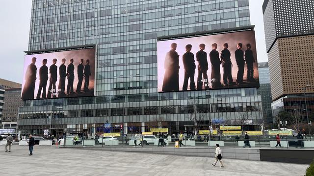 A modern building with a glass façade and two giant screens on it. The screens both display the same image: the 7 members of BTS standing in a line with foggy light around them. The street in front of the building is rather empty. 