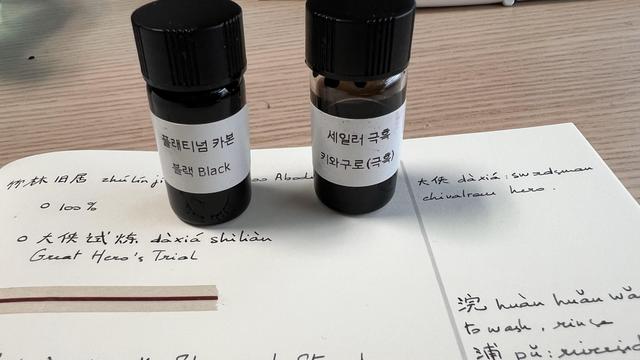 Two very small bottles of black ink with a label in Korean. They are on a notebook with various handwritten notes of Chinese vocabulary. 