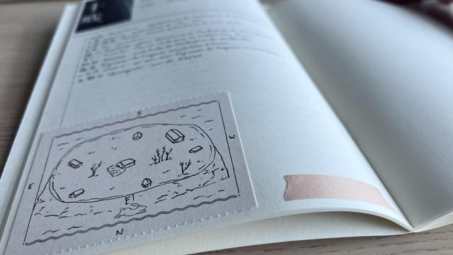 A notebook opened with the focus on the bottom half where a light cream memo sheet is glued. The drawing of an island with some isolated houses on it is on the memo sheet. The rest of the notebook page has some handwritten notes but they are out of focus.