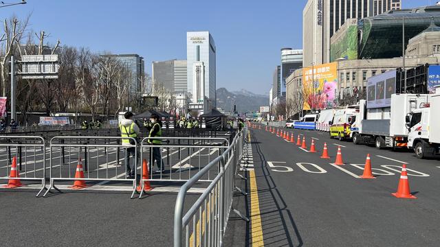 A large street lined with traffic cones and barricades and security agents in the middle. It’s closed to circulation and several vehicles are parked along the road. Tall buildings are visible in the background under a clear blue sky.