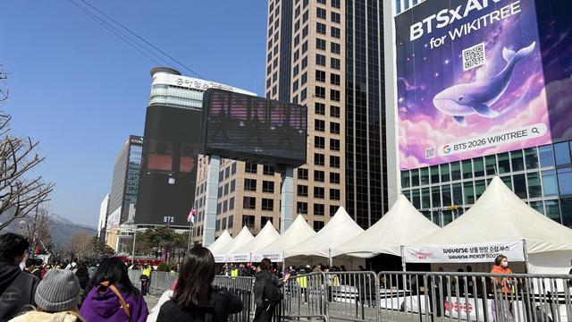 A city scene featuring tall buildings with giant screens and posters advertising for BTS. In front of the buildings there are booths to pick up goods. There are a lot of people walking on the sidewalk. 