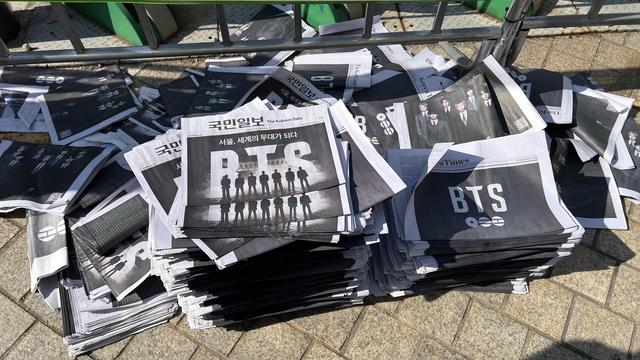 A pile of newspapers on the ground, outside. They are from different companies but all have a similar front page advertising for BTS with the name or pictures of the group featured prominently. 