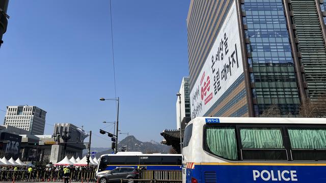 City scene with police buses jn the foreground and a tall building on the right with a huge banner on it featuring the Arirang logo. Booths and security officers can be seen in the background. 