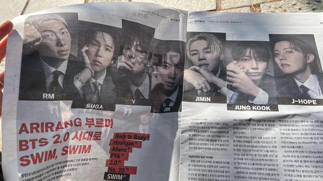 A newspaper held opened outdoor. It features pictures of the seven members of BTS and an article about the band and the album Arirang. 