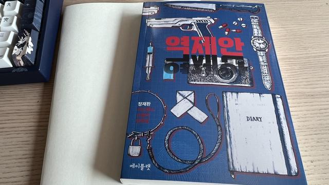A blue book in Korean is resting on a white notebook on a table. The corner of a keyboard is visible on the upper left corner. The book is the focus of the picture, it has the title 역제안 written in red and surrounded by ink drawings of objects like diary, watch, pistol, camera, knife, etc.