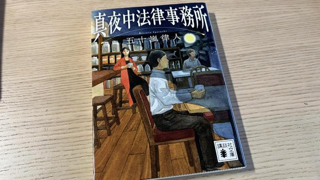 A book in Japanese on a wooden table. The cover displays a painting of three people in a coffeeshop. Two men are sitting at tables and looking towards the right, a woman is sitting at the counter and looking at us.