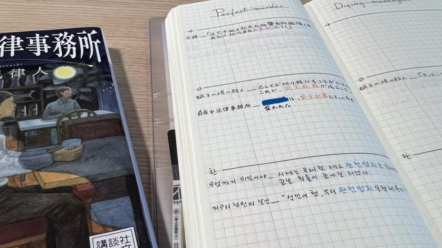 A long notebook with a focus on the left page. There’s the title “Perfect Murder” at the top and the page is divided into three sections, each containing some lines of text in different languages. A book in Japanese appears near the notebook, partly out of frame.